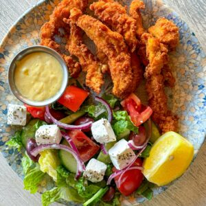 A plate of crispy fried chicken strips with Greek salad and dipping sauce.